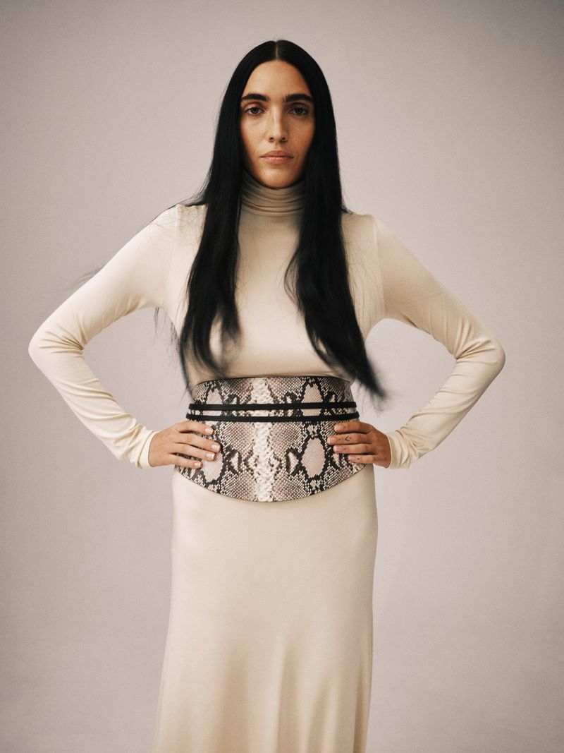 A slender adult woman poses indoors, wearing a long-sleeved, beige dress with a snakeskin-patterned belt and long, straight, black hair.