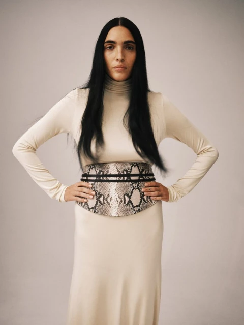 A slender adult woman poses indoors, wearing a long-sleeved, beige dress with a snakeskin-patterned belt and long, straight, black hair.