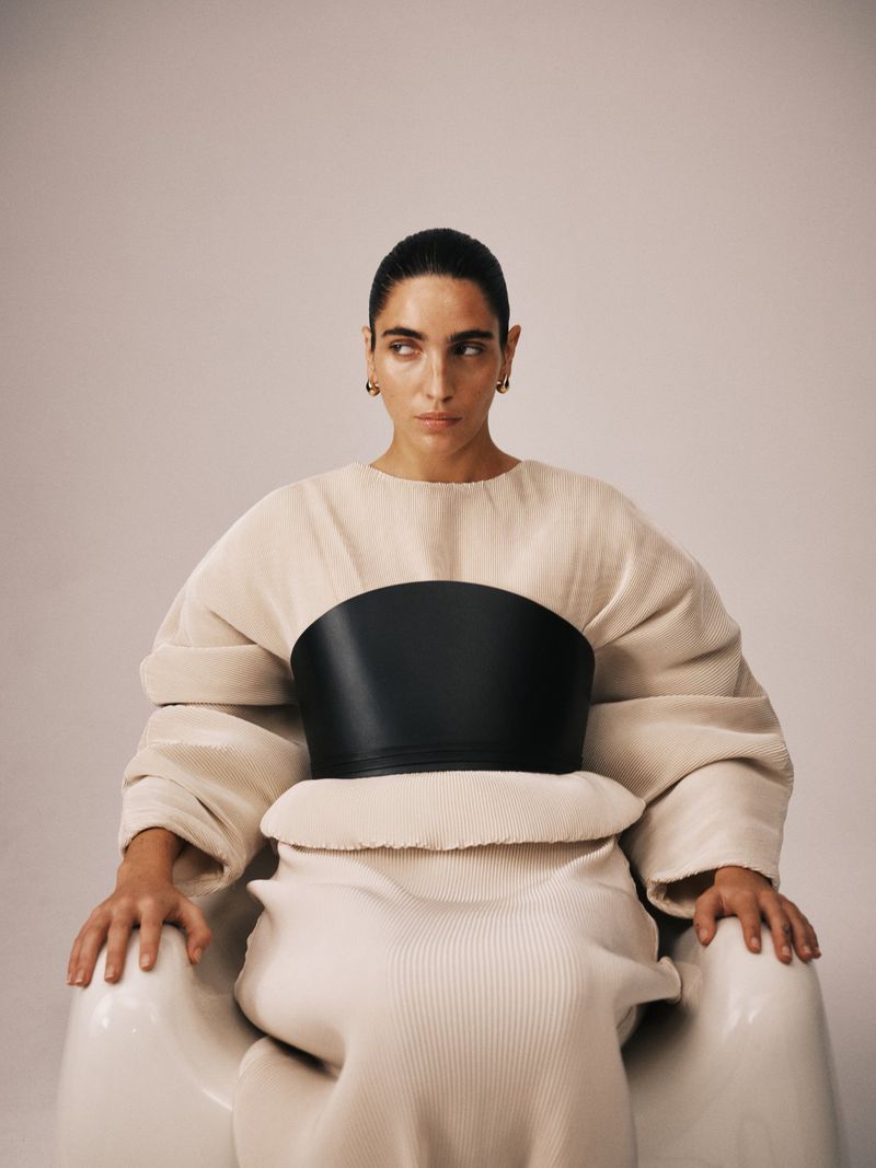 Woman seated wearing beige dress with a wide black leather belt around torso, staring off camera with focused expression. 