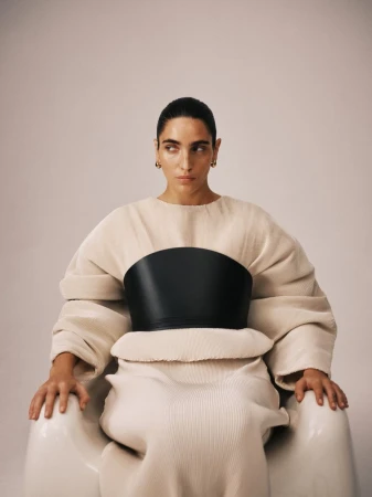 Woman seated wearing beige dress with a wide black leather belt around torso, staring off camera with focused expression. 