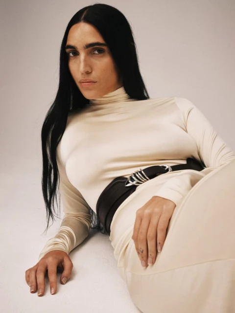 Woman in a beige dress with long dark hair laying on a beige background. She wears a wide black belt and has a neutral expression.