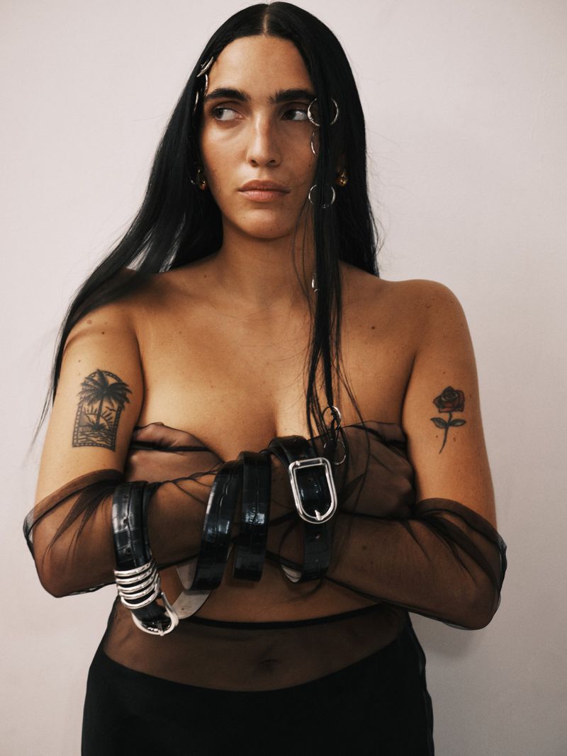 Adult female poses indoors with bare chest covered by belts and sheer fabric, accessories in hair, and tattoos on arms against a plain wall.