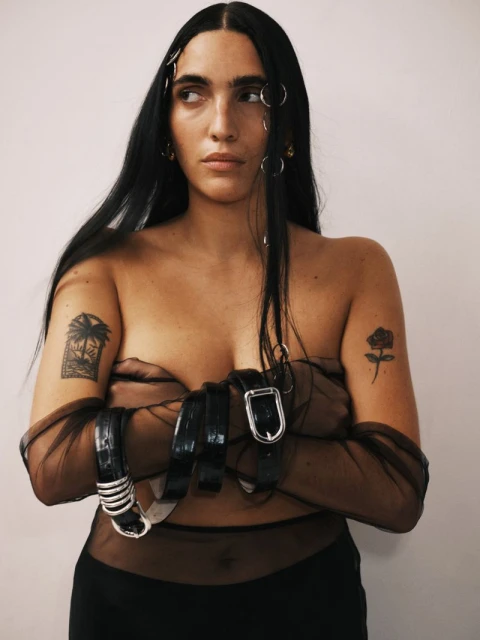 Adult female poses indoors with bare chest covered by belts and sheer fabric, accessories in hair, and tattoos on arms against a plain wall.