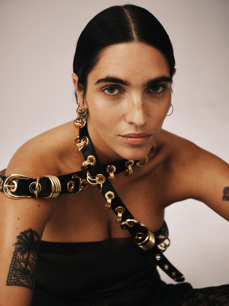 Portrait of a brunette adult female wearing a black outfit with gold hardware details in a studio setting with soft, neutral lighting.