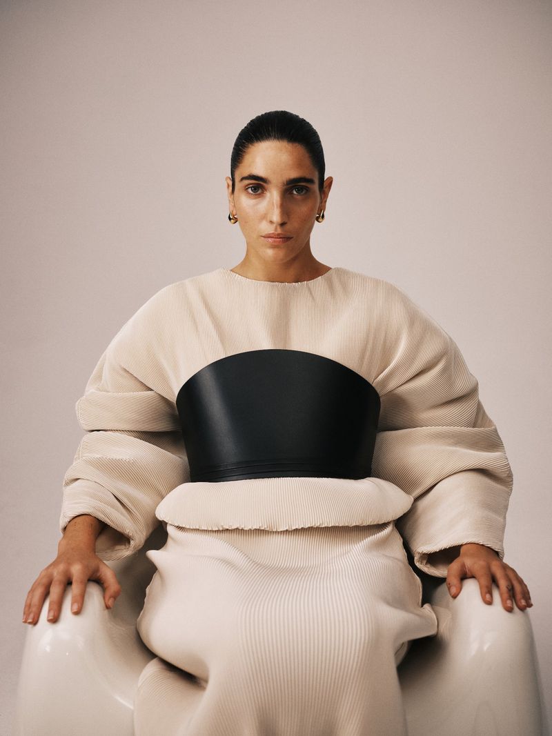 A woman in a flowing neutral-toned outfit and black obi sits in a modern armchair against a neutral backdrop, fashion photography.
