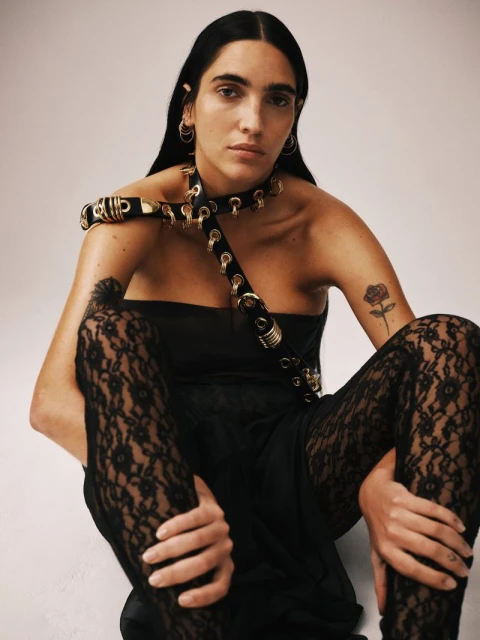 Woman in black dress and lace tights poses against white backdrop, wearing hoop earrings and harness-style necklace with gold accents.