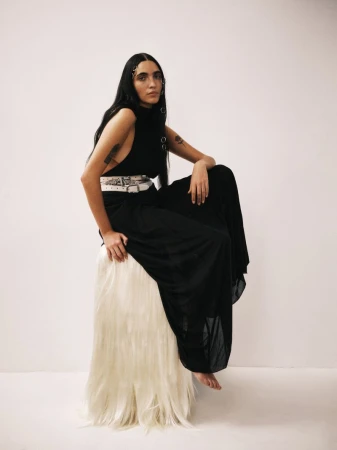 Model Valentine Valero seated wearing a black dress and layered white belts on a fringed plinth against a white seamless backdrop.