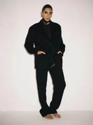 Model in a black suit and turtleneck with gold necklace standing against a plain white studio background