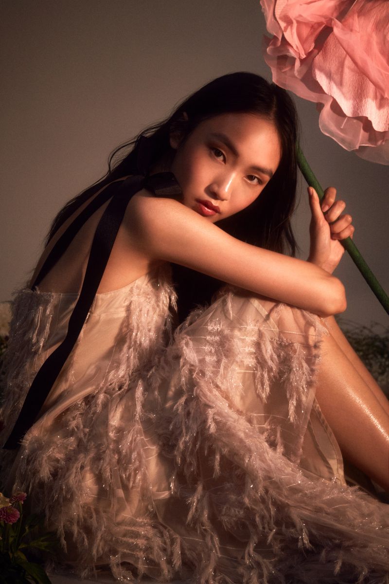 Asian woman in feathered dress poses seated beneath giant pink flower, captured in a studio with warm lighting and soft colors, editorial fashion shot.