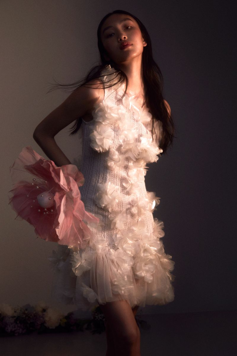 A stylish Asian woman in a white, feathery dress with a large pink floral accessory poses gracefully in a studio with soft lighting.