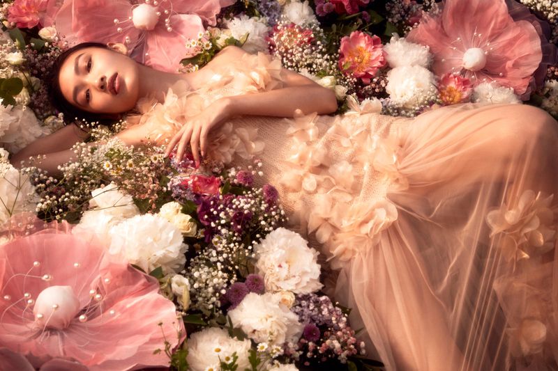 A beautiful Asian woman reclines amidst a vibrant array of colorful blossoms, dressed in a flowing pink gown that blends with floral background.