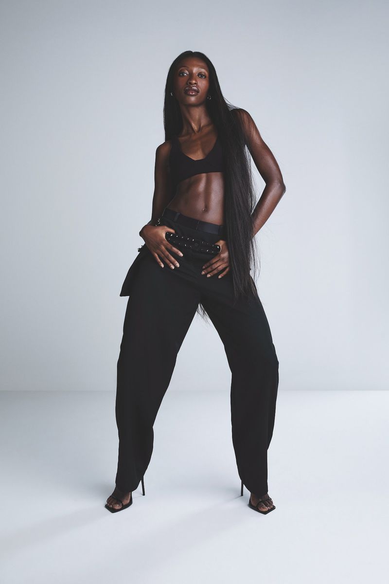 Elegant African-American woman posing in a black bra top and oversized pants against a plain backdrop in a fashion shot.