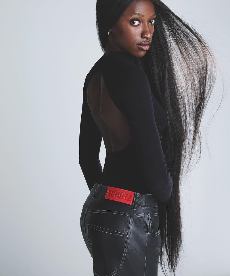 Fashion shot of a Black woman with long, flowing hair, wearing a black bodysuit and black pants, against a gray background.