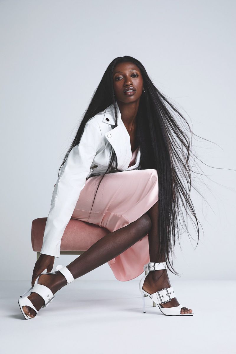 A Black fashion model sits on a pink stool wearing a white jacket, a pink skirt, and white heels against a white backdrop.