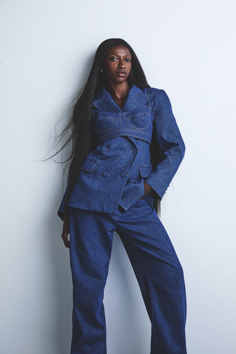 A black female fashion model poses in a double-breasted blue denim suit with long dark hair against a white backdrop in a studio shot.