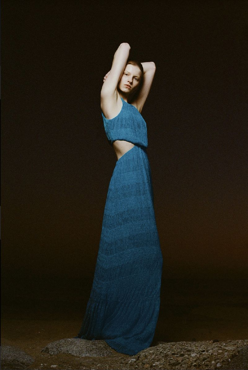 Fashion shot of a female model in a blue cut-out dress standing on rocks with her arms raised against a dark, neutral background.