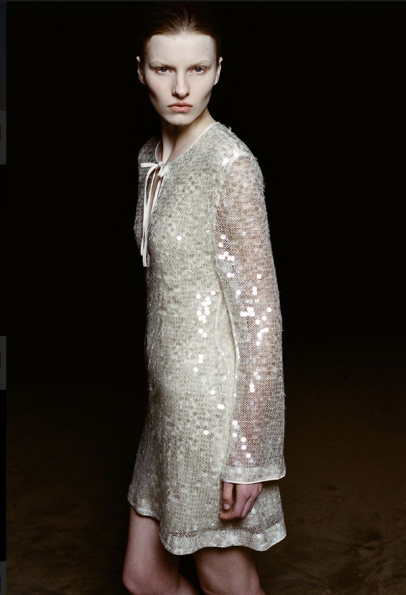 Fashion portrait of a pale-skinned woman with short hair, wearing a sparkly long-sleeved dress in front of a dark background.