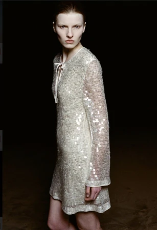 Fashion portrait of a pale-skinned woman with short hair, wearing a sparkly long-sleeved dress in front of a dark background.