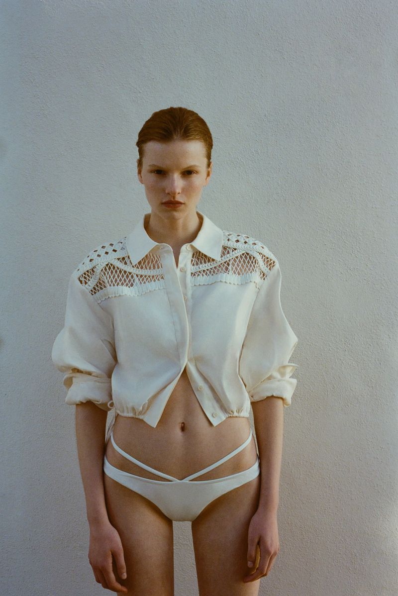 A woman poses in an open white button-down shirt and white bikini bottoms against a neutral wall in a studio shot. The image is brightly lit.