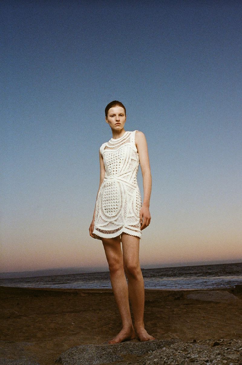 Fashion shot of a barefoot adult female wearing a white patterned dress outdoors on a beach with a blue sky at sunset or sunrise.