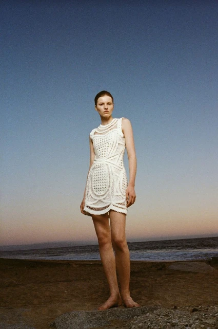 Fashion shot of a barefoot adult female wearing a white patterned dress outdoors on a beach with a blue sky at sunset or sunrise.