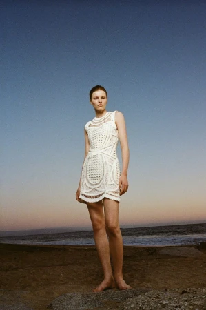 Fashion shot of a barefoot adult female wearing a white patterned dress outdoors on a beach with a blue sky at sunset or sunrise.