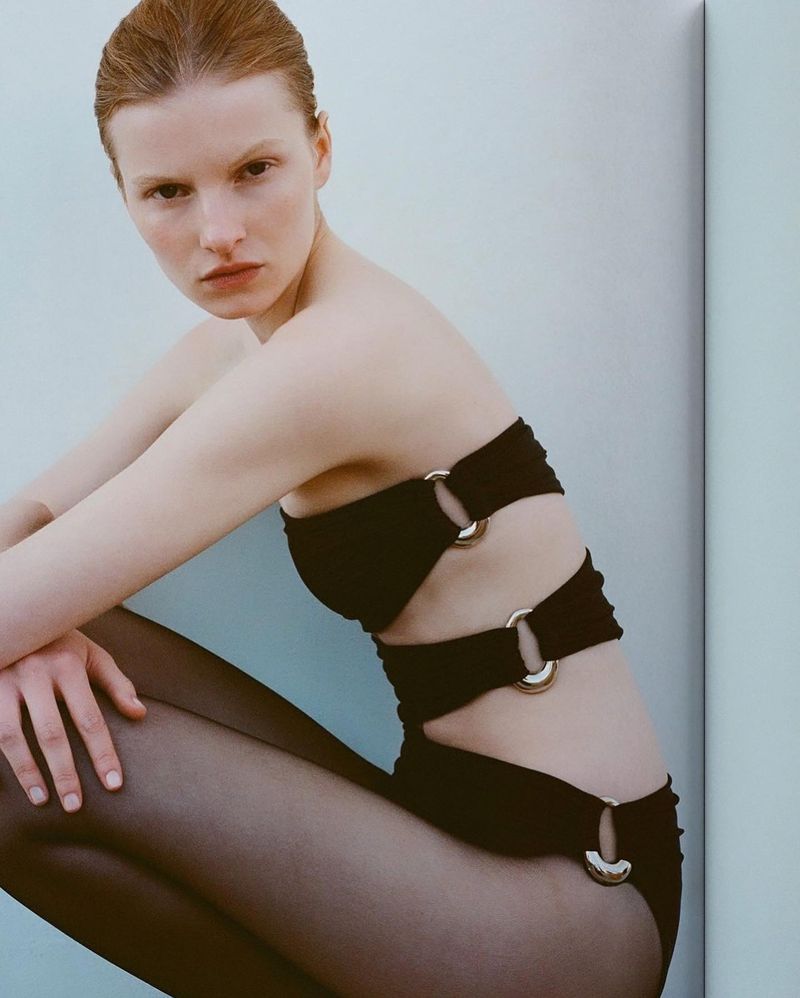 A woman poses in a black strapless swimsuit with gold rings, complemented by black pantyhose. She is indoors against a pale wall.