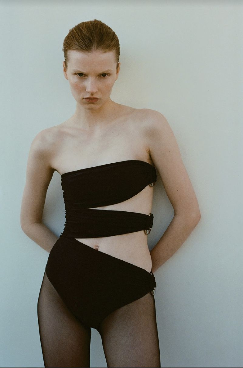 A solo adult female poses against a light wall, wearing a black strapless swimsuit with hip cut-outs and black fishnet stockings.