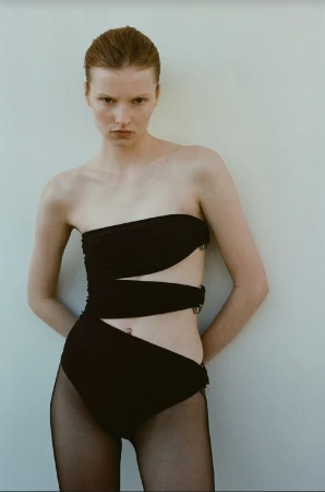 A solo adult female poses against a light wall, wearing a black strapless swimsuit with hip cut-outs and black fishnet stockings.