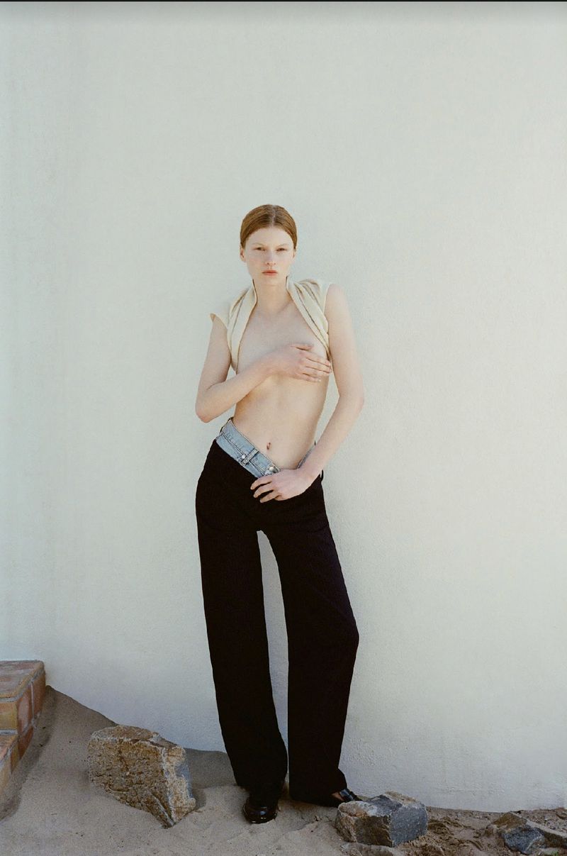 Full-length studio shot of a topless, fair-skinned adult female with auburn hair wearing black pants with a silver belt, against a white wall.