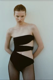 Model Lainey Hearn in a strapless black cutout bodysuit and sheer black tights against a plain studio background for Simkhai.