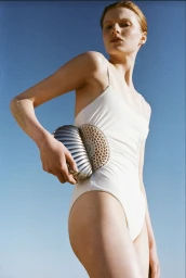 Model wearing a white one-piece swimsuit and holding a metallic shell handbag against a clear blue sky.