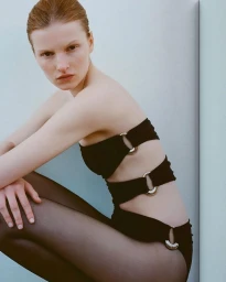 Model Lainey Hearn crouched in a strapless black cutout swimsuit with metallic ring hardware and sheer black tights.