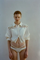 Model wearing a cream-colored buttoned shirt with cutout shoulders and matching bikini bottoms against a plain textured wall