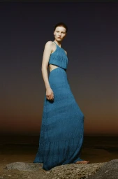Model standing on rocky ground wearing a blue cut-out maxi dress against an evening sky background.