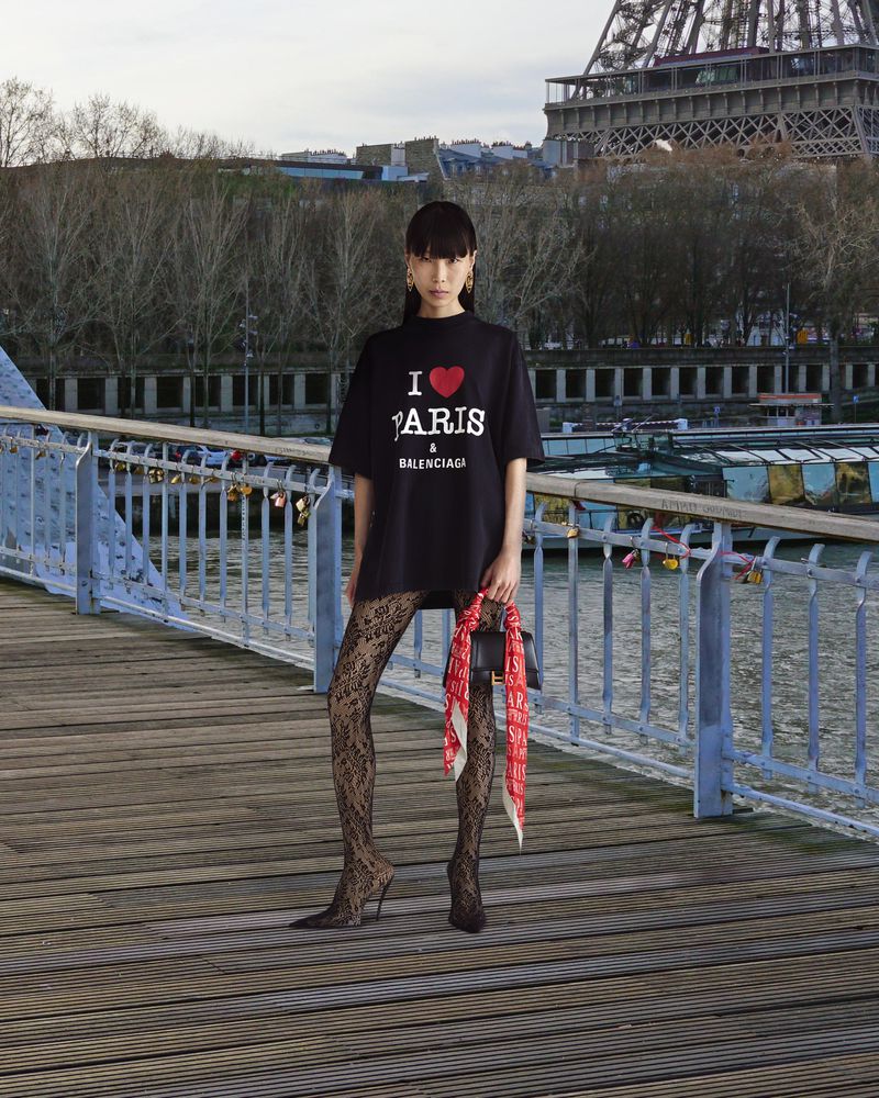 Woman posing in front of Eiffel Tower wearing 'I Love Paris' t-shirt, patterned tights, holding purse with scarf in Paris, France.