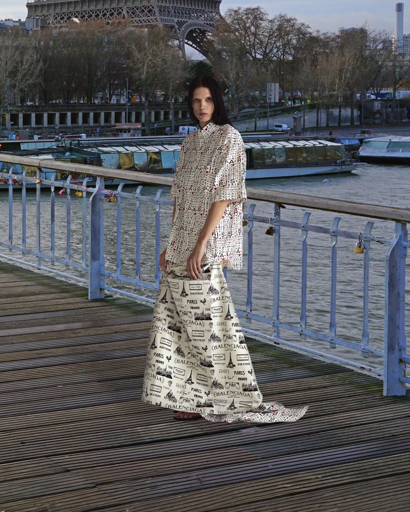 Fashion shot of a woman in a patterned skirt and top, standing on a bridge with the Eiffel Tower and boats in the background.