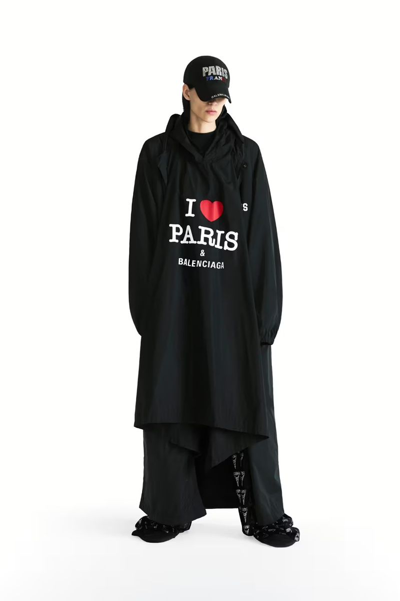 Full-length studio shot of a model wearing a black Balenciaga hooded jacket with 'I Love Paris' print, paired with black pants and a cap.