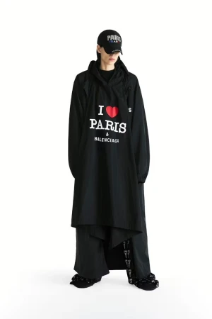 Full-length studio shot of a model wearing a black Balenciaga hooded jacket with 'I Love Paris' print, paired with black pants and a cap.