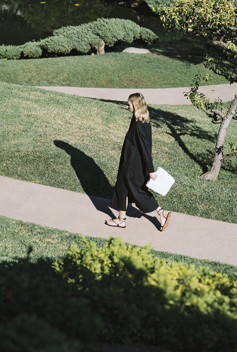 A woman in a black dress is walking on a path in a park with green grass and trees. She is carrying a white object.