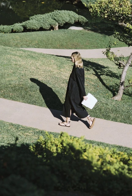 A woman in a black dress is walking on a path in a park with green grass and trees. She is carrying a white object.