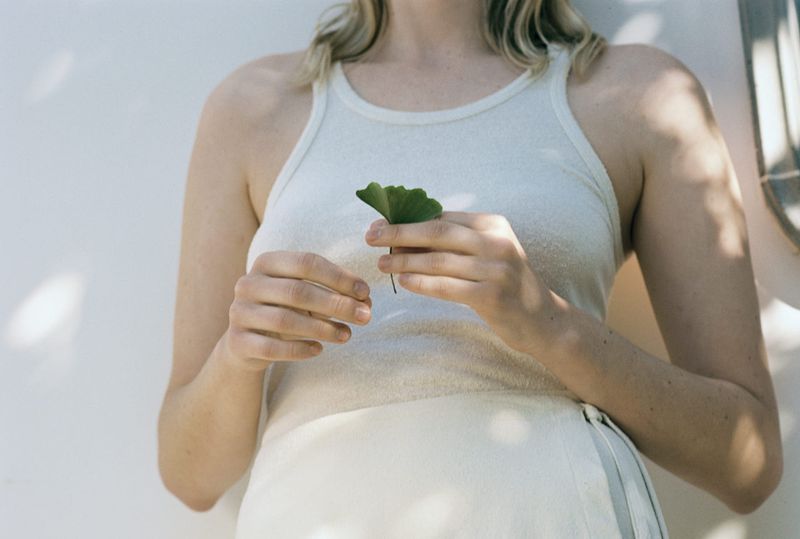 Blonde woman holds green ginkgo leaf in soft light, wearing white tank top and skirt, standing against a simple white background.