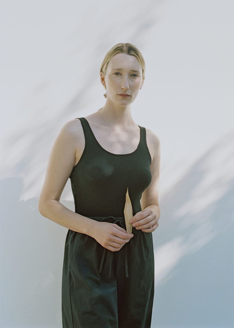 Woman in dark tank top and pants holds a wooden stick against a white wall with shadows. Simple portrait showcases casual style.