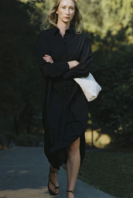 Blonde woman in a black shirt dress walks on an outdoor path with a white bag, sunlight, and dark foliage.