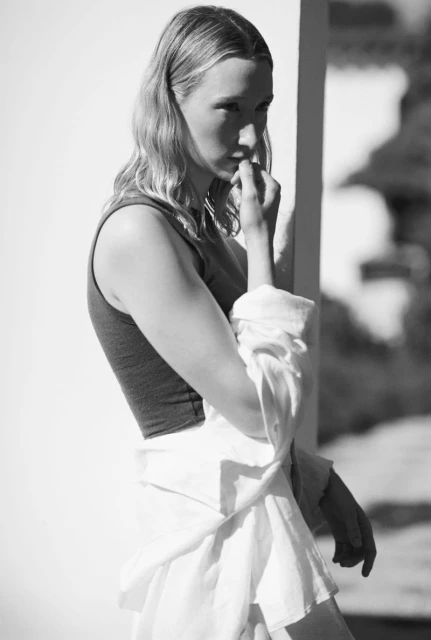 Black and white portrait of a blonde adult woman in a tank top with a white shirt tied around her waist, hand near her face.