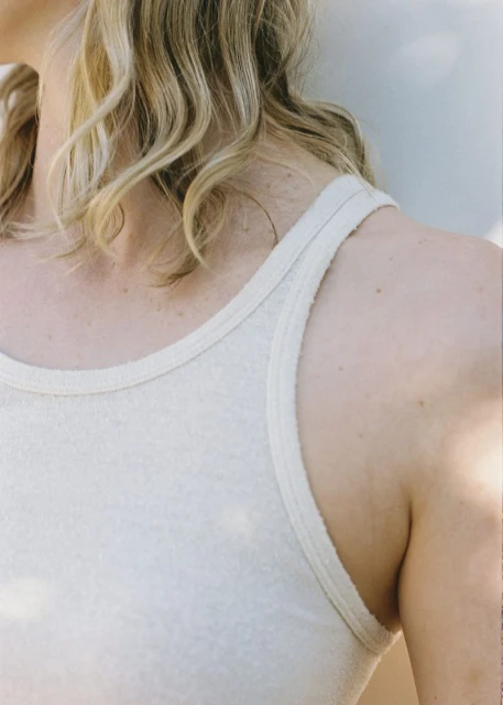 Close-up of a fair-skinned woman with wavy blonde hair wearing a light-colored tank top against a neutral background in soft, natural light.