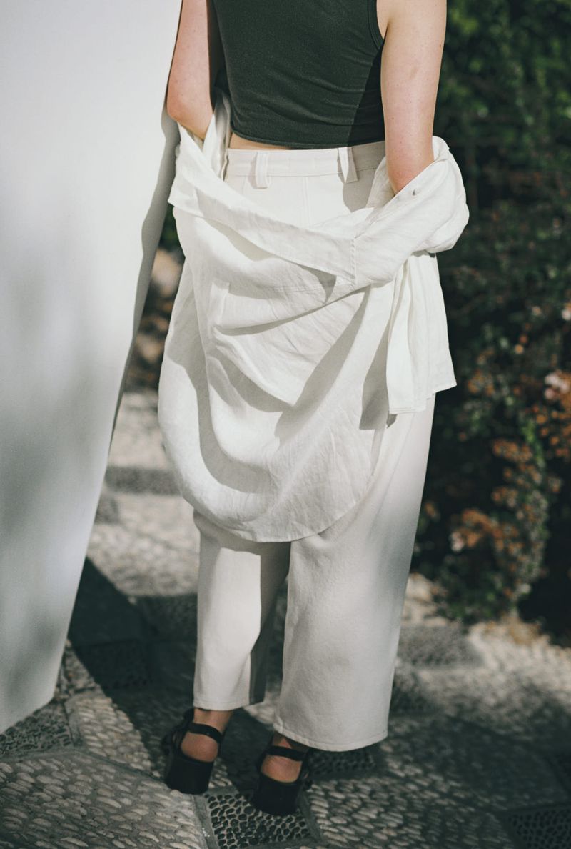 Back view of an adult female in a dark green tank top and white pants, wearing black sandals and an unbuttoned white shirt.