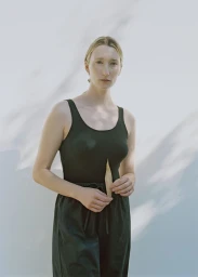 Model in dark green tank top and matching trousers holding a leaf against a white wall with leaf shadows