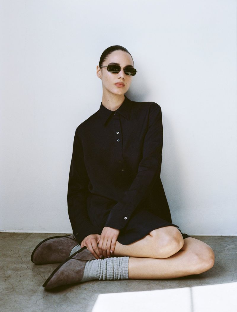 A young woman in black dress and sunglasses sits on the floor in front of a white wall, fashion photography, studio lighting, medium shot.