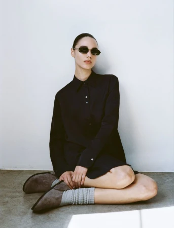 A young woman in black dress and sunglasses sits on the floor in front of a white wall, fashion photography, studio lighting, medium shot.